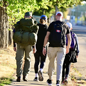 Image shows two people walking. One is wearing a black hoodie with a Canadian flag and the other is wearing a military uniform carrying a ruck sack.