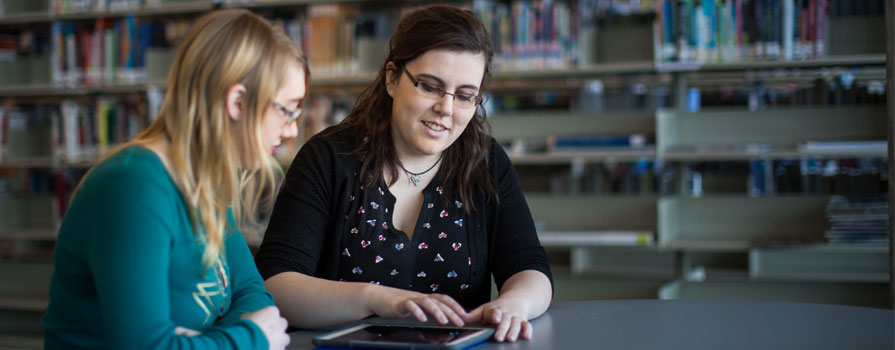 Two individuals sit at a desk in a library. One person is assisting the other by showing them something on an iPad.