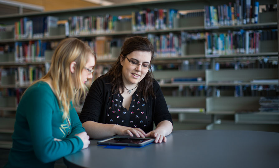 Two individuals sit at a desk in a library. One person is assisting the other by showing them something on an iPad.
