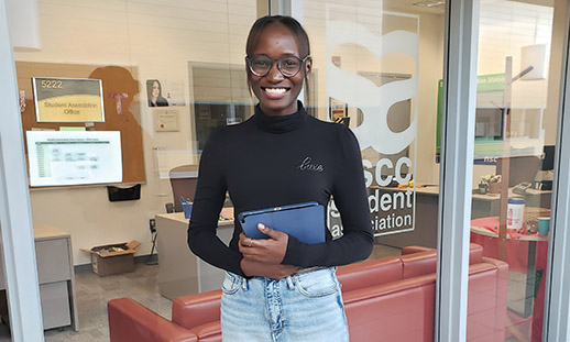 Ribi holds a binder in front of her and stands in front of the Student Association office at Ivany Campus.