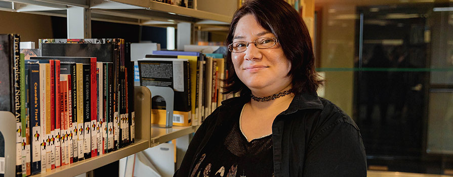 Riki Lee Christmas is shown holding a book titled “The Encyclopedia of the First Peoples of North America.” She is wearing a black, long sleeve shirt and a choker-style necklace. She is wearing glasses.