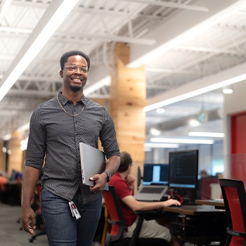 Sandrico Provo walks through an office space smiling and holding a laptop computer.