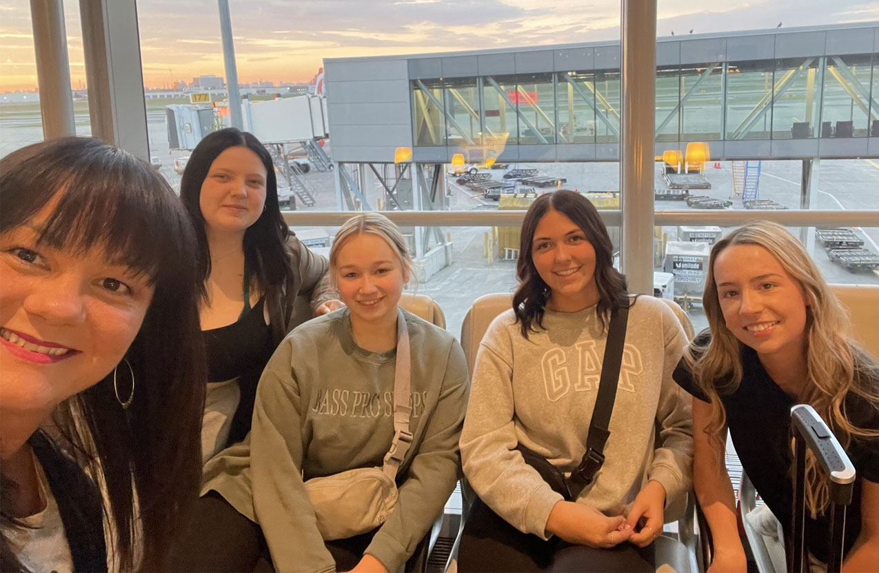 Five people take a photo together at the airport with the sunrise behind them.