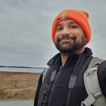 Mitchell stands outdoors near the ocean wearing a hat, coat and backpack.