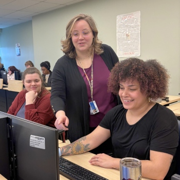 Students are working on a computer with a faculty member pointing to the screen.