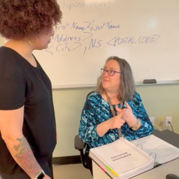 A student is talking to their teacher in a classroom setting. The teacher has a large binder open on the desk.