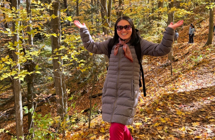 Image shows a person outside in the woods, smiling with their hands up. 