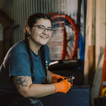 Taylor O'Hearn wears glasses, a t-shirt and safety gloves. She kneels in a workshop in front of a tool box and smiles at the camera.