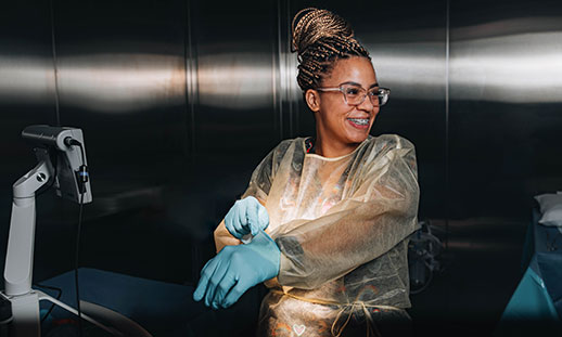 Vanity Thompson, a nurse, smiles while looking off to the side. She is wearing a yellow surgery gown and putting on a blue, surgical glove. She is in a stainless-steel room.