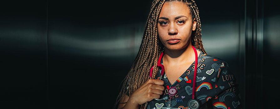 Vanity Thompson, a nurse, looks seriously at the camera. She is wearing a red stethoscope around her neck. Her scrubs feature rainbows and hearts. She is in a stainless-steel room.