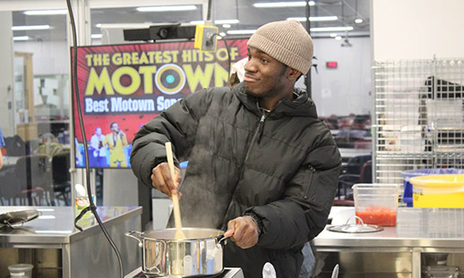 Wearing a black puffer coat, Victor stirs a large stock pot in an industrial kitchen.