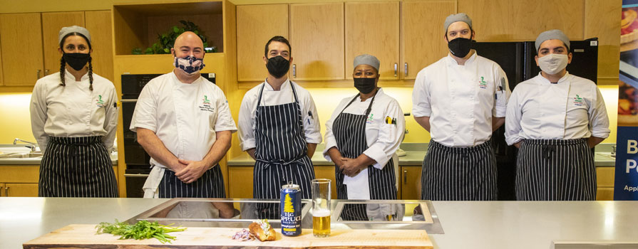 Six chefs wearing uniforms and facemasks stand behind a long counter. There are cupboards behind them. On the counter is a can of beer, a glass of beer and some produce.