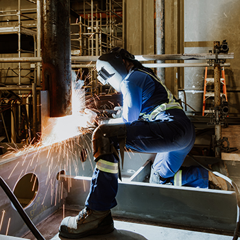A person wearing coveralls and a welding helmet kneels on their right knee and welds.