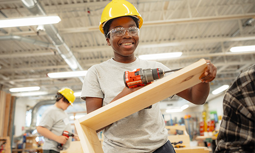 Youth in carpentry shop holding a piece of wood from a project while smiling