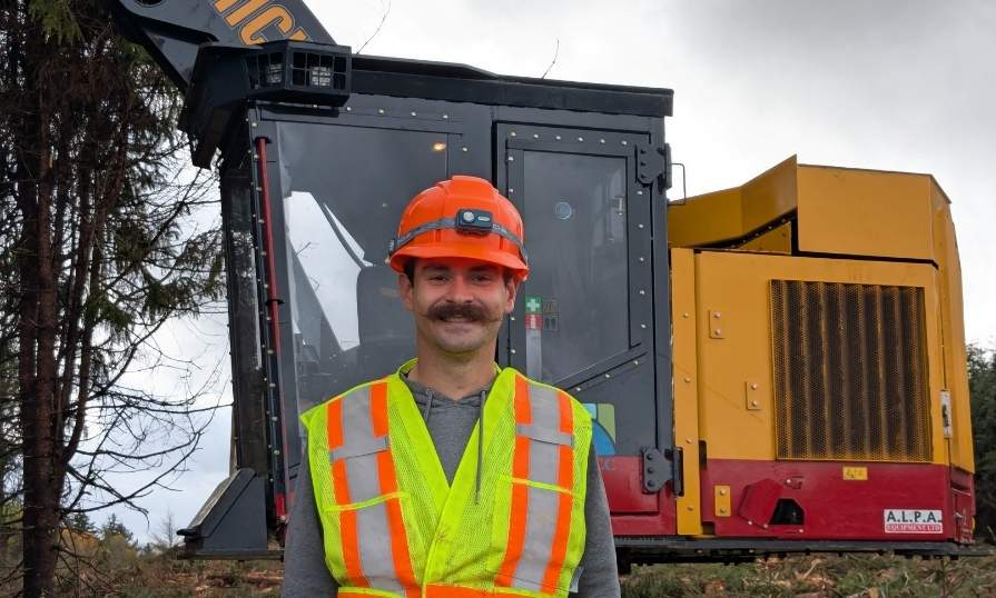 A person outdoors wearing a safety helmet and vest smiling in front of forest machinery. 