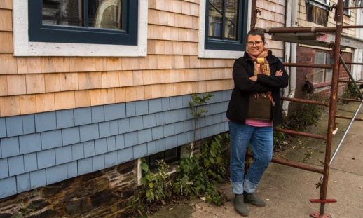 Image shows a person standing outside a building that is under construction with their arms folded across their chest.