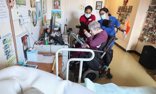 Two CCA's assist a patient that is in a wheelchair, providing care to the individual in their room.