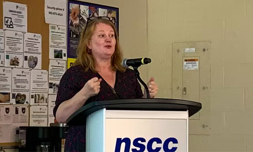A person stands at an NSCC-branded podium, speaking to an audience.