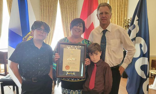 A woman and her two young sons stand in the Lieutenant-Governors office with him, receiving a framed award.