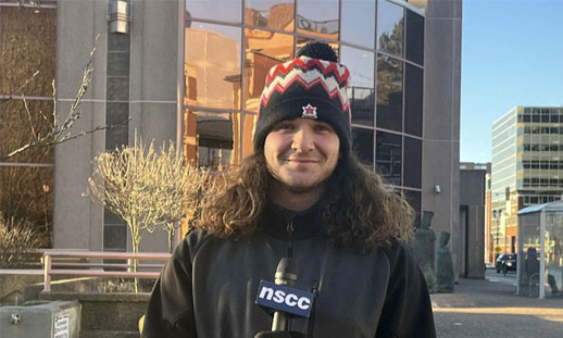 A male with long dark hair wears a black and red hat, gloves and a jacket while standing outside a building with a 'NSCC' branded microphone in hand, prepared to do an interview.
