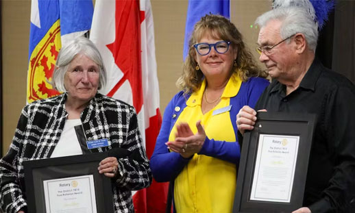 Three individuals stand together, two holding framed certificates they'd just been awarded. 