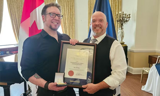Two individuals pose for a photo in the Lieutenant-Governor's office while holding a framed award.