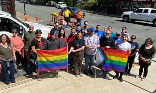 2SLGBTQ+ allies stand as a group on the sidewalk with Pride flags in hand.