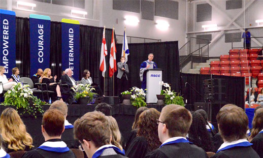 The camera faces the stage, as an official addressees the crowd at a graduation ceremony.