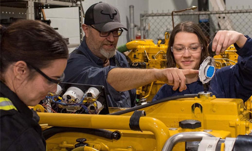 A male and two females work on a yellow piece of marine machinery.