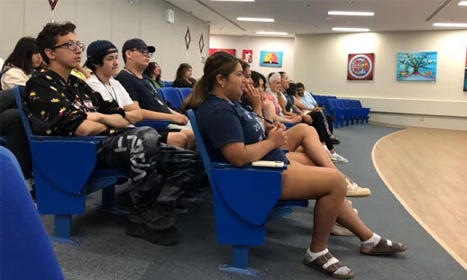 Youth are sitting in blue chairs during a presentation in an auditorium.