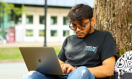 Person wearing a black shirt that says "here we code", sitting outside under a tree looking at a laptop
