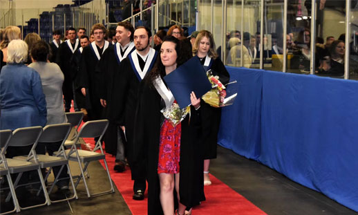 A line of graduates walk down a red carpet with their diplomas in hand, exiting the graduation ceremony. 
