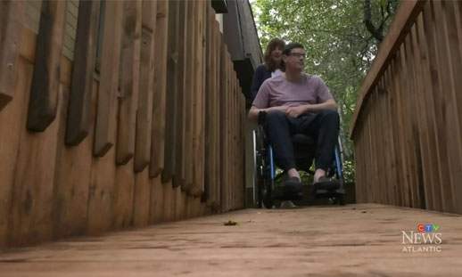 A male in a wheelchair is shown being transported by his mother across a new, wooden ramp outside his house. 