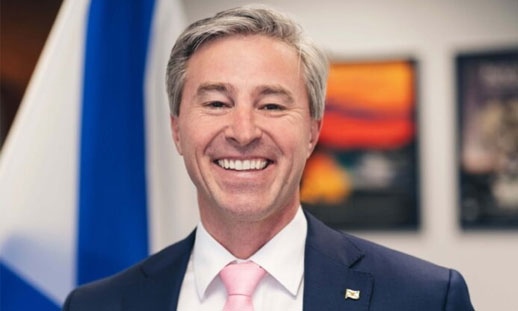 A man is smiling. He has grey hair, a black suite, white dress shirt and pink tie on. The Nova Scotian flag is in the background.