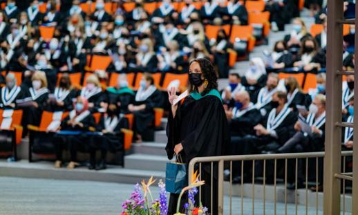 Woman in convocation robe wearing a mask and waving in front of an auditorium of fellow graduates.