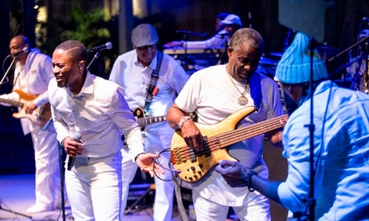 Five people are on stage, singing and playing musical instruments while all wearing white outfits.