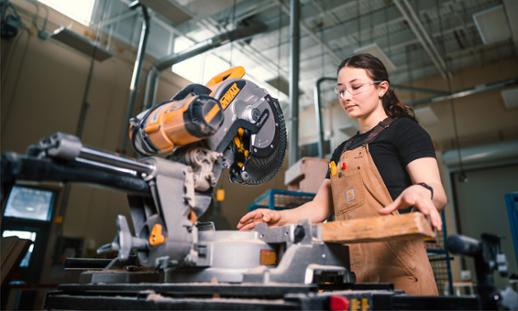 A female carpentry student prepares a piece of wood before cutting it. She is wearing clear safety glasses and brown Carhart overalls with some tools in her front pocket.