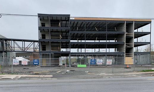 The metal framing of a new building is under construction. The weather is cloudy and the construction site is fenced in.
