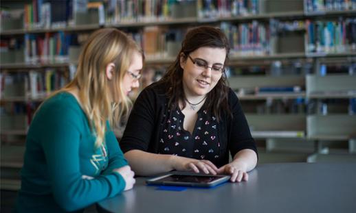 Two individuals sit at a desk in a library. One person is assisting the other by showing them something on an iPad.