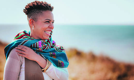 A woman wearing a colourful scarf, smiles broadly while looking towards the horizon. Water can be seen in the background.