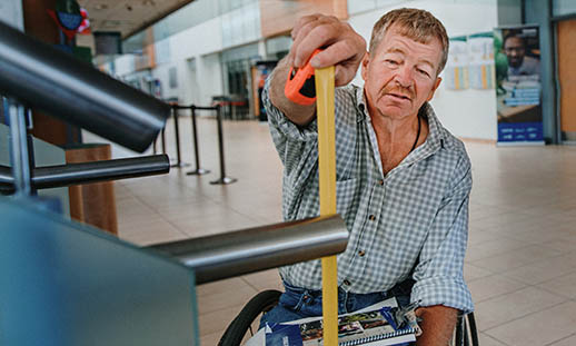 A man sitting in a wheelchair and wearing a buttoned-up plaid shirt and jeans, measures a railing with a yellow measuring tape. 