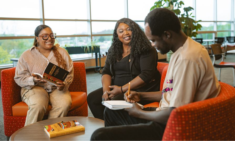 Image shows 2 NSCC students and their faculty sitting in a library setting with books and notepads.