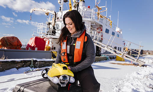 A woman with brown hair is crouched down. She is wearing an inflatable live-vest and holding a small underwater drone that is yellow. In the backgroun, a large, white ship and a gangway can be seen.