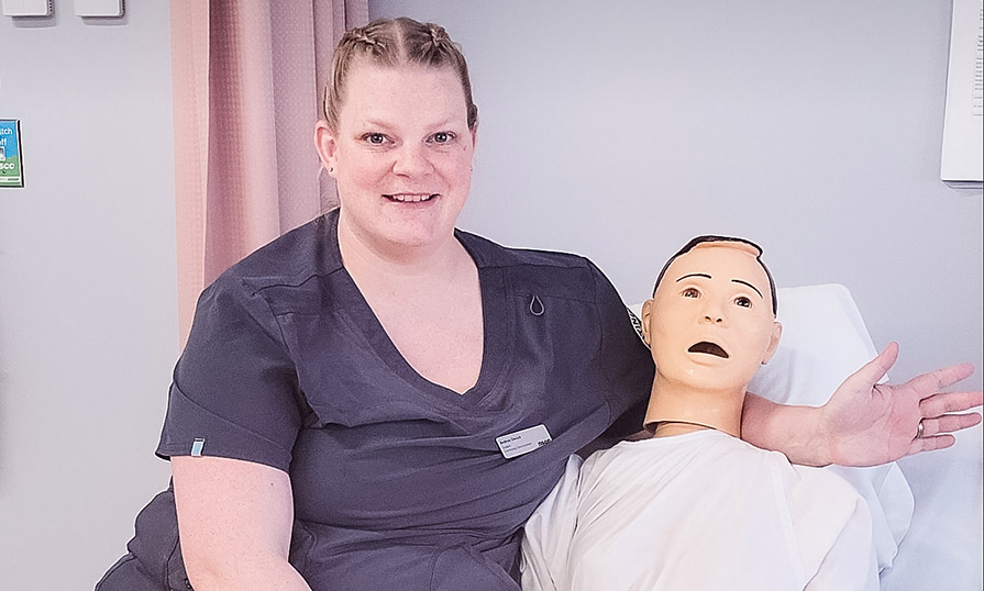 Andrea sits on a hospital bed beside a male mannequin wearing a hospital gown.