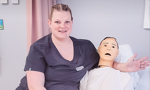 Andrea sits on a hospital bed next to a male mannequin wearing a hospital robe.