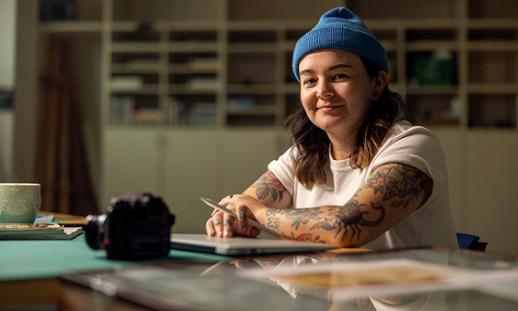 Anisa Francoeur sits at a table. She wears a t-shirt, beanie hat and holds a pencil while smiling at the camera.