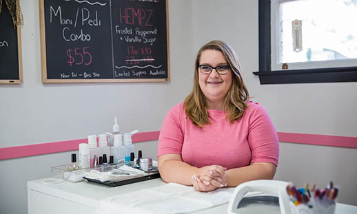 "A young woman in a pink shirt sits behind a desk and smiles. On the desk, nail polish and other aesthetic equipment is seen. There is a chalkboard in the background that reads mani/pedi combo. "