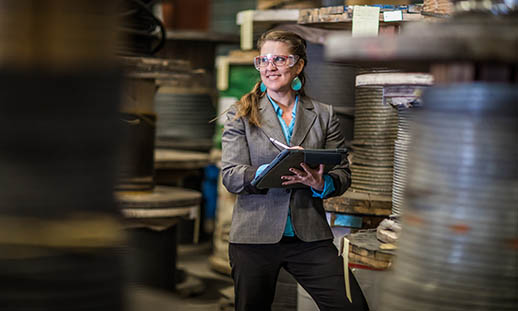 A woman wearing a blazer, safety glasses and large, turquoise earrings can be seen through a frame made by industrial ropes and supplies. She is looking off to the side of the image and writing on a clipboard.