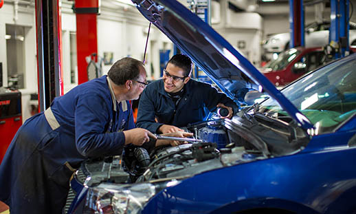 Two men talk while leaning over an open car engine bay. The car and both men's workwear is also blue. They are in an automotive shop.