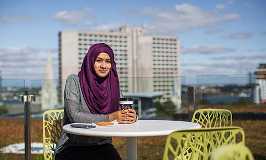 A woman, wearing a purple hijab sits outdoors in a modern green chair at a small round table. In the background tall buildings can be seen. 